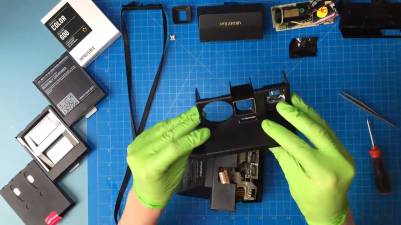 A Person Wearing Green Gloves Disassembling a Polaroid Camera with Tools and Film Packs Nearby